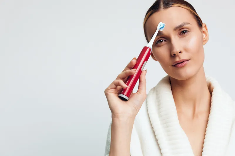 Young woman holds electric toothbrush near face with gentle expression wearing white bathrobe isolated on light background healthy dental care concept hygiene routine.