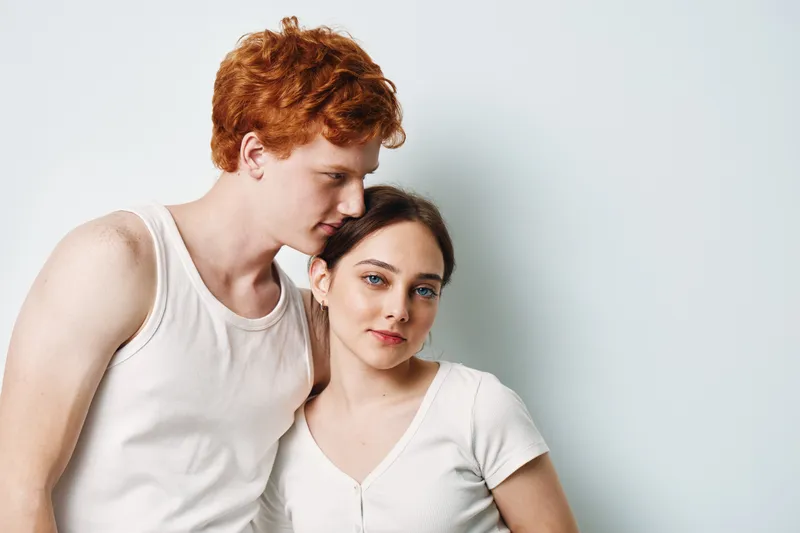Young couple posing together with neutral expressions against white background. Man with red hair and woman with blue eyes standing close in casual white clothing portrait.