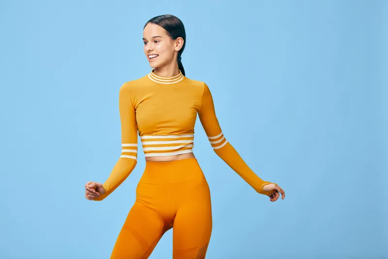 Woman fitness sport: young woman fitness model in orange sportswear crop top and leggings with long sleeves smiles and poses in a studio against a smooth blue background.
