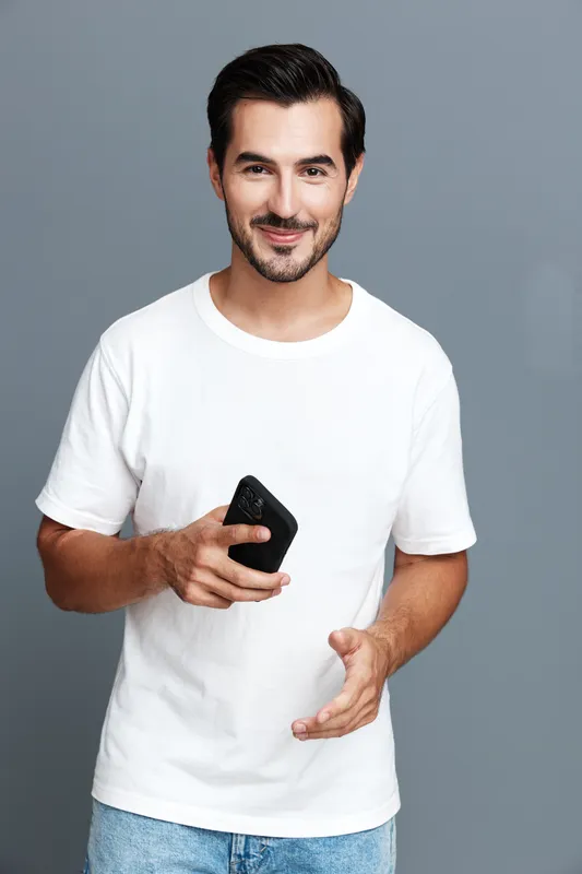 Smiling man holding smartphone, wearing casual white t-shirt, looking at camera, posed against a minimalist gray background. People lifestyle concept
