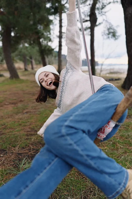 Girl on a swing in an outdoor park, laughing with joy as she soars, cozy sweater and jeans, trees in background, candid moment of carefree childhood play
