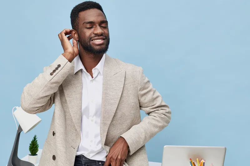 Professional young African American man in beige blazer and white shirt, smiling with closed eyes while listening to music with earphones, standing at modern workspace with blue background, studio por