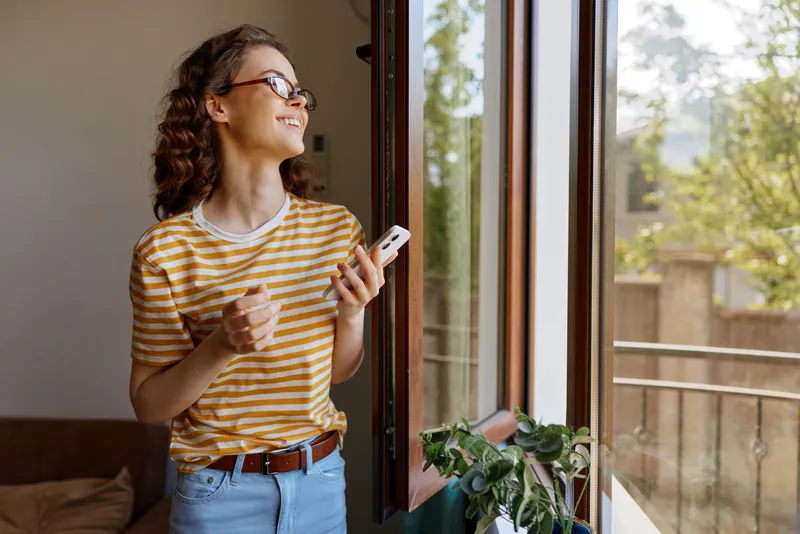 Casual woman indoors by a sunlit window, smiling while holding a smartphone, natural daylight, cozy home atmosphere, relaxed lifestyle moment, warm tones, cheerful mood