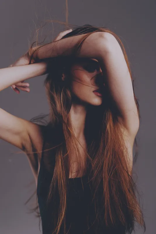Artistic woman with long hair covering face in a moody studio setting with soft lighting and muted colors