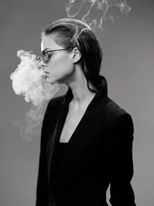 Black and white smoking woman with glasses in dark jacket exhaling smoke in studio