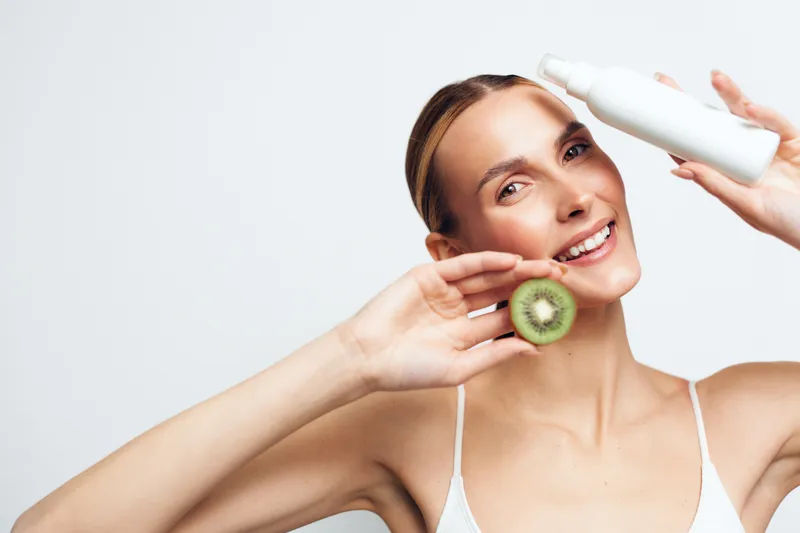 Woman skincare session, holding kiwi and applying cream on her face, smiling with relaxed posture, soft studio lighting, clean white background, fresh beauty ritual for skin care