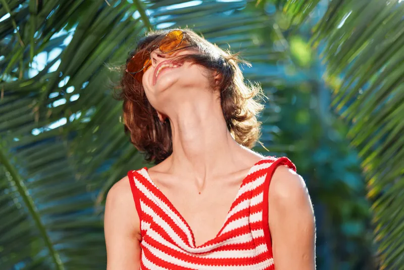 A cheerful woman basks in sunny outdoor light, tilting her head back while wearing a red and white striped top, surrounded by lush palm leaves, conveying carefree summer vibes.