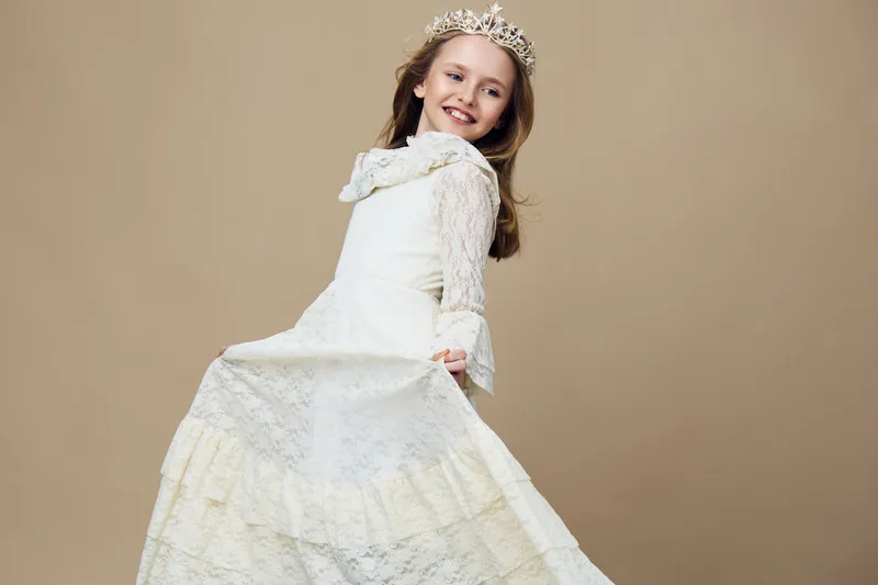 Young girl in elegant white dress with a crown, joyful expression, posing against a soft beige background, evoking innocence and charm, ideal for children s fashion themes