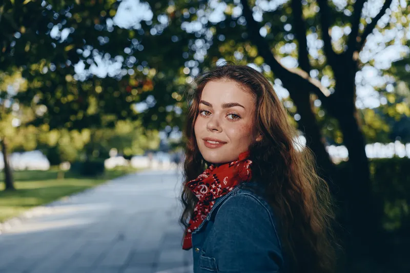 smiling woman with freckles and curly hair wearing denim jacket and red scarf outdoors in sunny park with green trees background