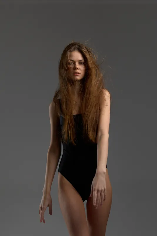 Woman with long hair in black bodysuit posing confidently against gray background with soft lighting.