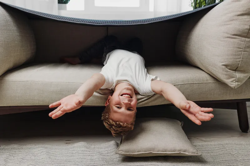 Playful boy with curly hair upside down under couch stretches arms forward in cozy living room. Child having fun, carefree moment, and indoor play concept with natural light and home comfort.
