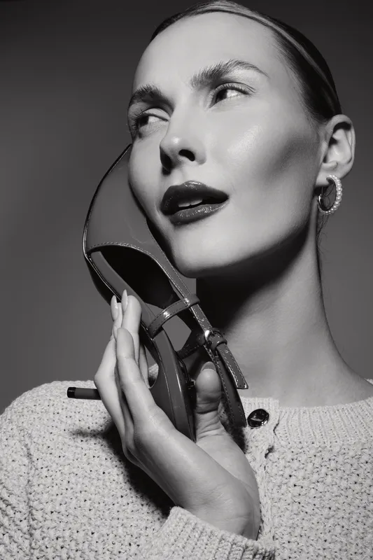 Portrait of a young white woman with pale skin holding a high heel shoe near her face in black and white studio lighting.