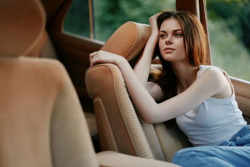 A young woman in casual wear relaxes in a car's backseat, showcasing a serene expression and natural beauty in a soft, warm color palette.