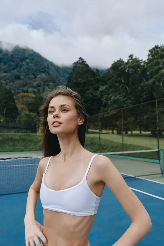 Young athletic woman in a white crop top poses confidently on a tennis court, capturing an energetic and sporty vibe amidst a scenic background