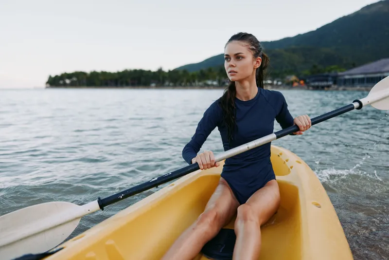 Young woman paddling a bright yellow kayak on calm water, gripping a double-bladed paddle with focus. She wears a navy long-sleeve top, hair tied back, immersed in a scenic lakeside view.