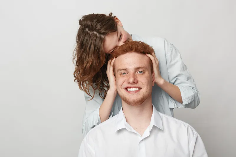Happy young man with red hair smiles while woman with curly hair hugs him from behind on grey background. Concept of friendship, affection, and positive emotions.