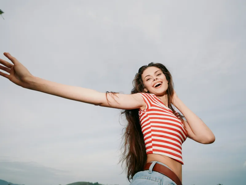 Joyful young woman with long hair smiles widely, arms extended, wearing a striped crop top and denim shorts, set against a bright sky, conveying carefree summer energy.