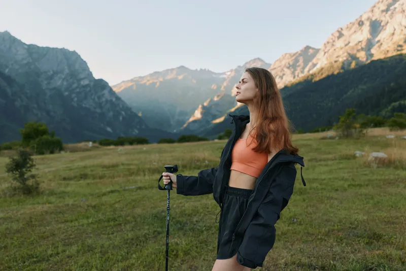 A young woman stands outdoors in a mountain meadow, wearing a windbreaker and athletic gear, gazing into the distance with a calm, determined expression as rugged peaks rise behind.
