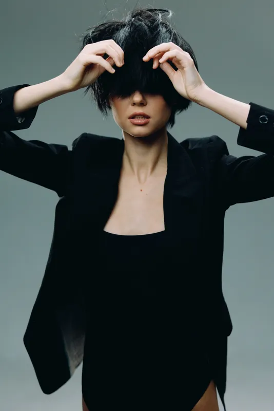 Stylish woman in black outfit lifting hair with hands, modern fashion, confident pose, minimalistic background, beauty portrait.