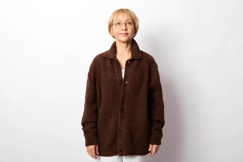 Elderly woman with glasses, studio portrait, wearing warm brown sweater jacket, calm friendly expression, standing on white background, soft evenly lit light