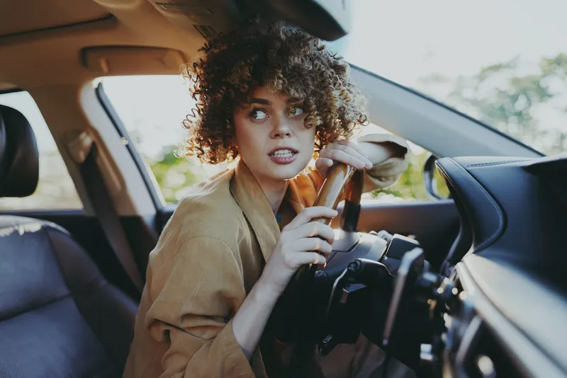 A young woman with curly hair sits behind the steering wheel in a car, looking back with a wary, focused expression during a casual city drive, capturing hesitation.