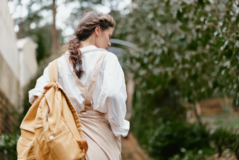 Young woman in casual outfit walking in nature, wearing a backpack and a white shirt with suspenders, evoking a sense of adventure and freedom in a lush green environment