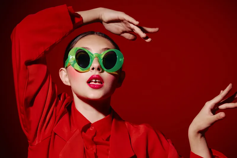 Fashionable young woman with glossy red lips poses wearing oversized green sunglasses and a vibrant red outfit. High contrast studio shot on red background, creative beauty concept.