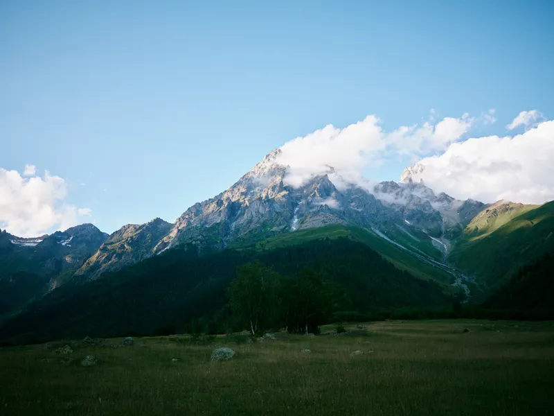 Mountains rise over a green meadow and distant forest under a blue sky with clouds, a tranquil nature landscape ideal for outdoor exploration and travel inspiration for picture use