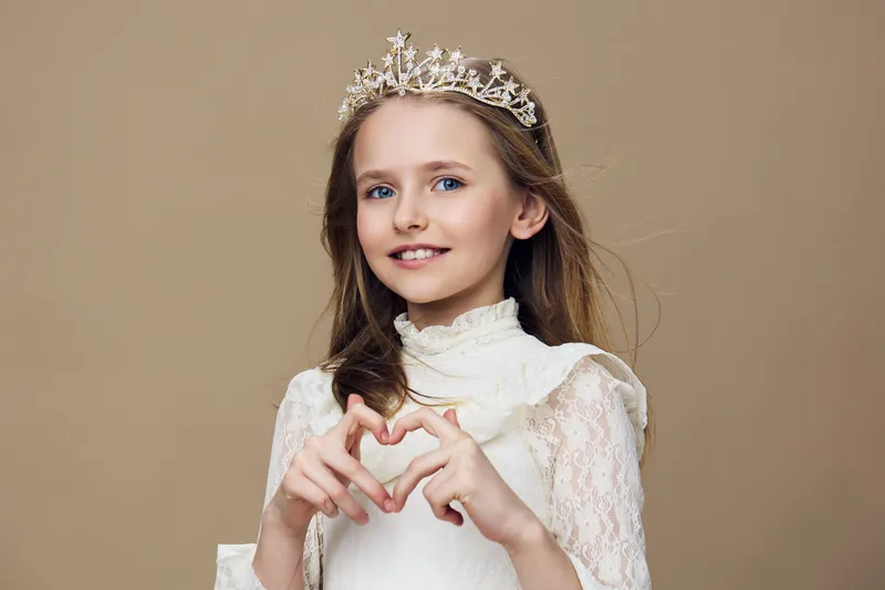 Cute girl with a tiara forming a heart shape with her hands, dressed in a white dress, smiling against a beige background, perfect for children s photography
