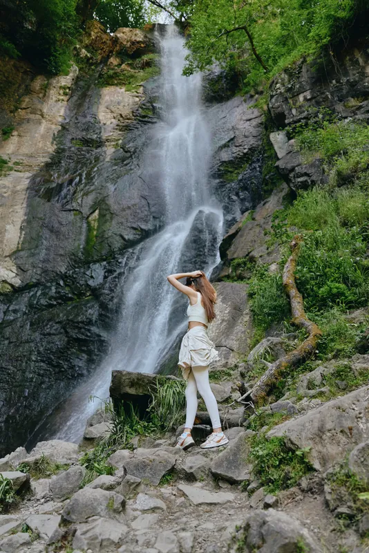 Woman hiking near waterfall in lush green forest, wearing sporty white outfit, enjoying nature and summer outdoor adventure with rocky terrain background.