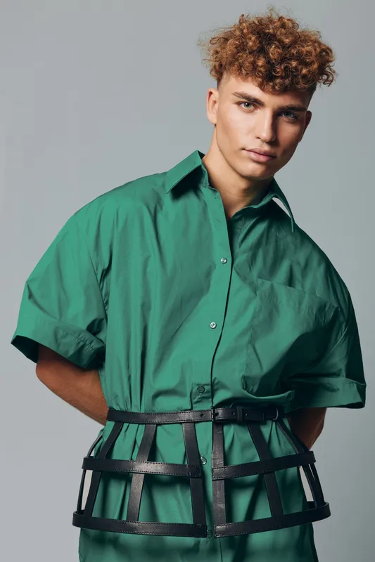 Young man with curly hair wearing green oversized shirt and black cage belt posing against gray background in studio. Fashion model with serious expression and modern style clothing.