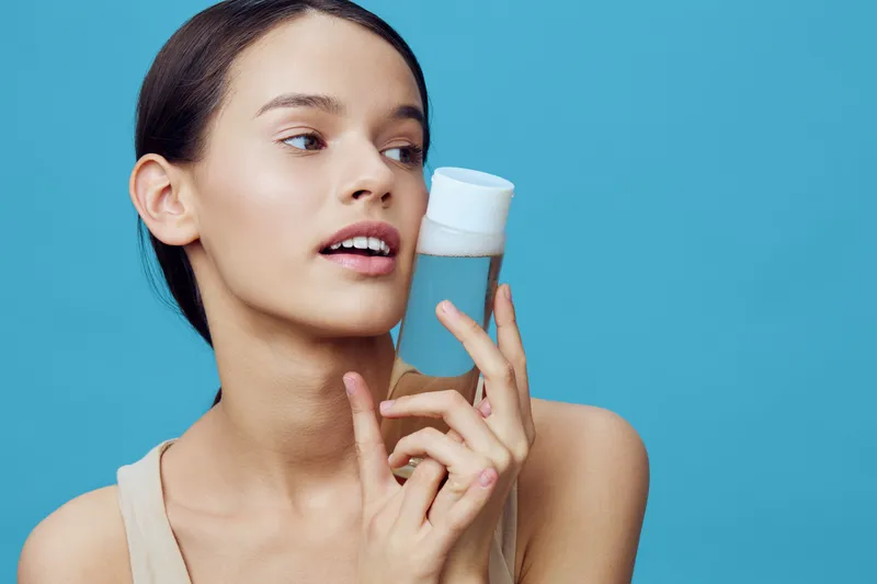 Young woman with natural beauty holding a skincare bottle, showcasing a fresh and radiant look against a blue background Ideal for beauty and skincare themes
