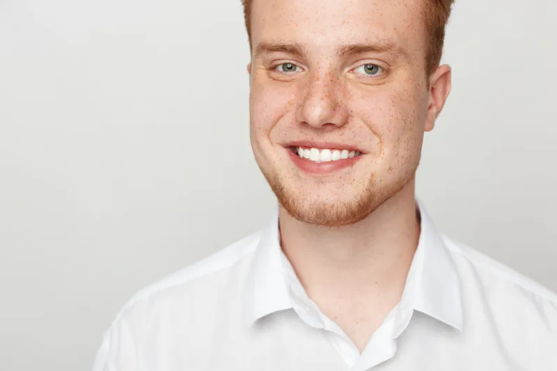 Smiling young man with red hair and beard wearing white shirt looks at camera. Close-up portrait on light background. Concept of happiness, confidence, and casual style.