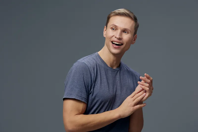Portrait of a smiling man with light brown hair wearing a casual blue t-shirt, standing against a plain gray background. Happy, cheerful expression, casual pose, male model, lifestyle concept.