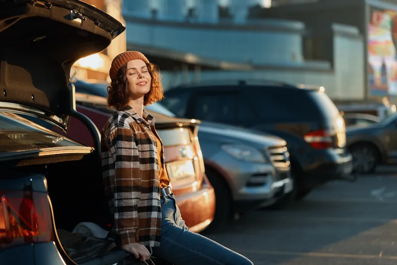 Woman by car trunk in parking lot at sunset, smile and beanie, plaid jacket, candid lifestyle and authenticity with golden hour glow, mindful living and emotional storytelling vibe.