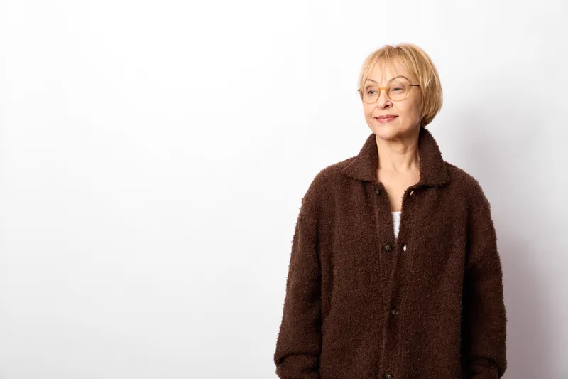Senior woman portrait, smiling and wearing glasses, brown coat on white studio background, calm friendly expression, half body framing, copy space for text, indoors.