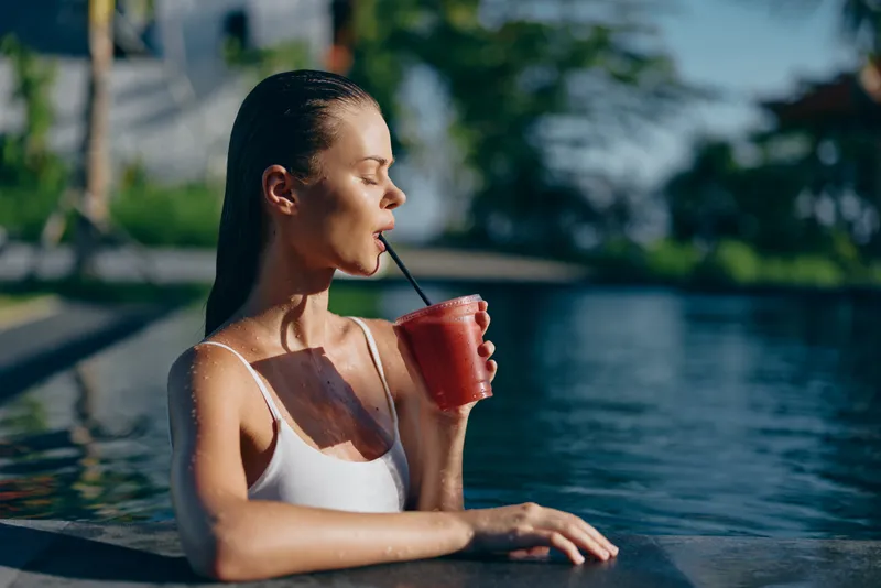 Relaxed woman lounges in a pool, sipping a red smoothie through a straw, wearing a white swimsuit, sunlit water backdrop, serene summer vibes, casual leisure by the poolside, bright holiday mood.