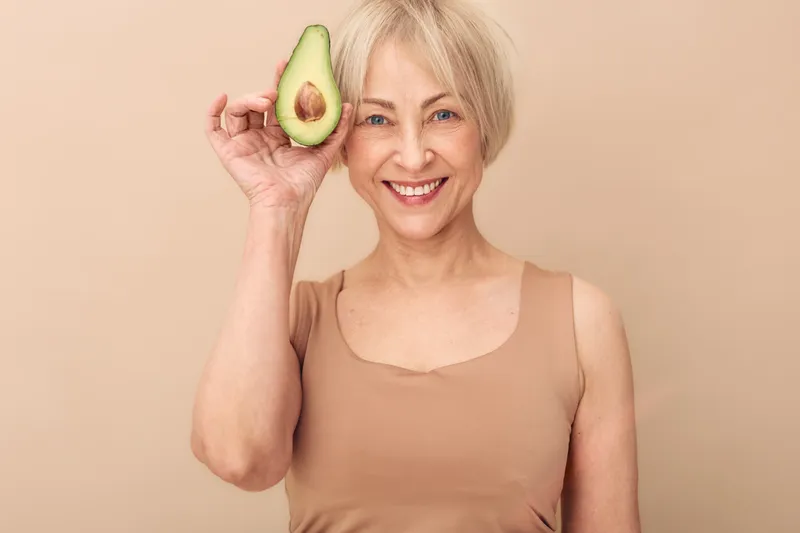 Smiling mature woman in beige top holds half an avocado near her face against a neutral background. Concept of healthy lifestyle, nutrition, wellness, and natural beauty for seniors.