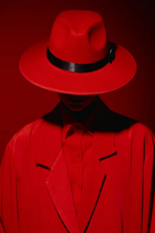 Portrait of a mysterious model wearing a red wide-brimmed hat and bright red coat, partially obscuring their face in dramatic red studio lighting. Creative fashion beauty concept.