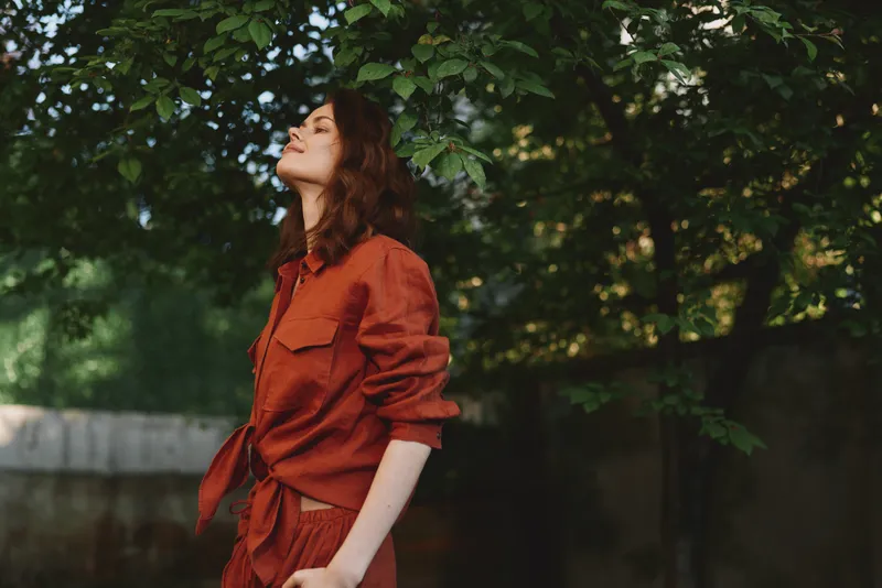 A woman in a rust red outfit stands outdoors among green leaves, eyes closed, head tilted back, savoring a calm moment, embodying mindfulness and nature
