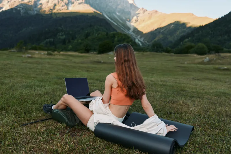 A woman sits on a grassy meadow with a laptop, facing a distant mountain range. She smiles softly, enjoying a relaxed and productive outdoor work moment.