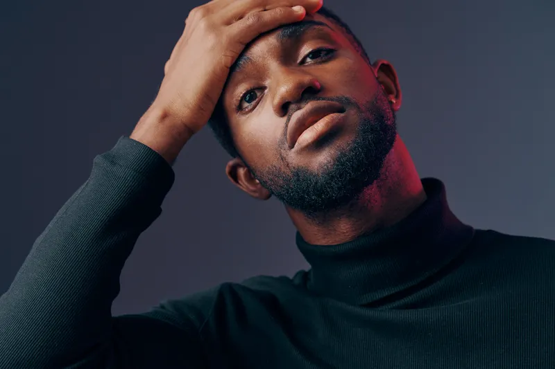 A confident young man dressed in a black turtleneck sweater poses thoughtfully against a dark gradient background, showcasing a modern and expressive look