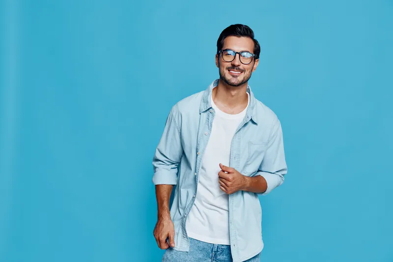 Smiling man wearing glasses and casual outfit standing against a blue background, looking at the camera with a friendly expression, showcasing a relaxed vibe.