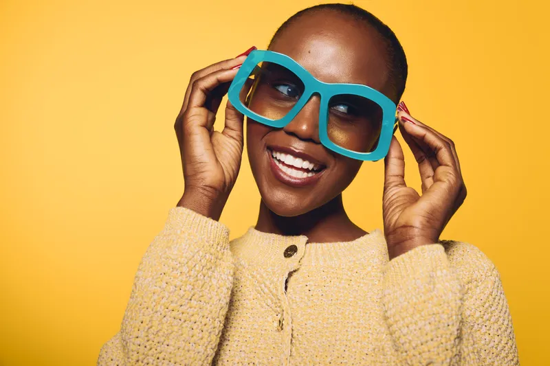 Happy dark skinned African woman wearing oversized blue sunglasses and a beige sweater smiles joyfully against yellow background. Concept of fashion, beauty, and positive mood.