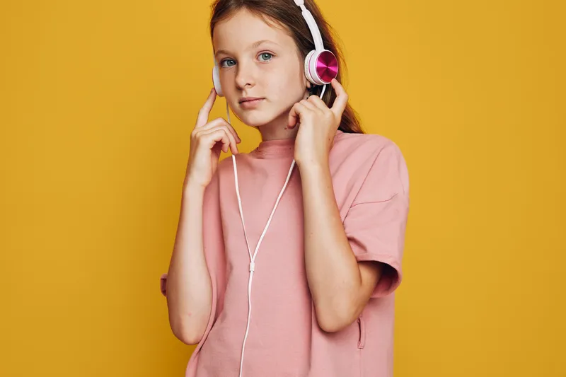 Young girl in a pink sweater listens to music with headphones against an orange background. She looks calm and focused with a neutral expression.