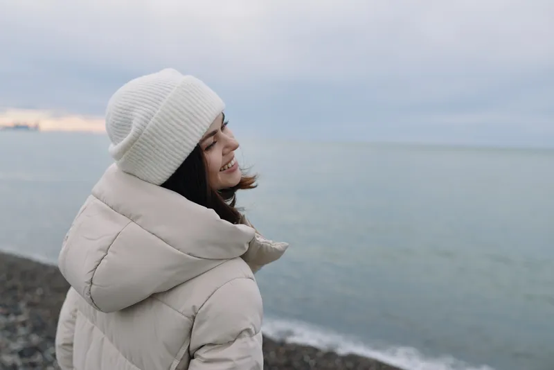 Warm winter scene featuring a woman in a white beanie and puffy coat by a calm coastline, smiling as the sea meets the cloudy horizon.