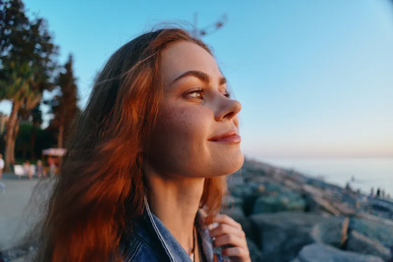 lifestyle woman in modern boho-western denim street style with natural makeup, warm film color, enjoying serene outdoor sunset by rocky shore.