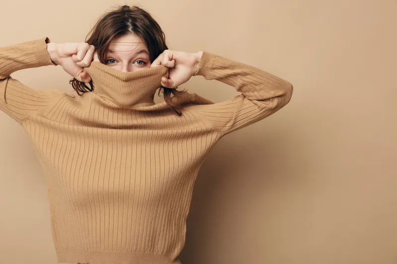 Young woman in beige knitted sweater pulls collar over face, peeking with eyes. Studio portrait on neutral background. Concept of cozy fashion, autumn style, and playful mood.