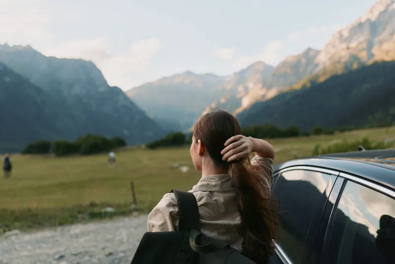 Young woman with long hair stands by a car in a mountain valley, looking at the distant peaks during a calm late afternoon.
