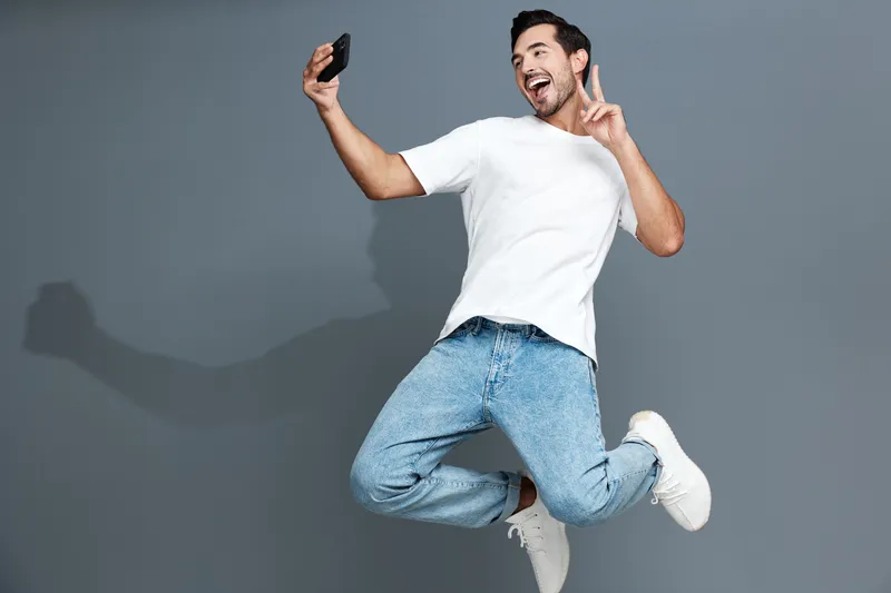 Joyful young man jumping in the air while taking a selfie with his smartphone against a gray background, having fun and expressing happiness.